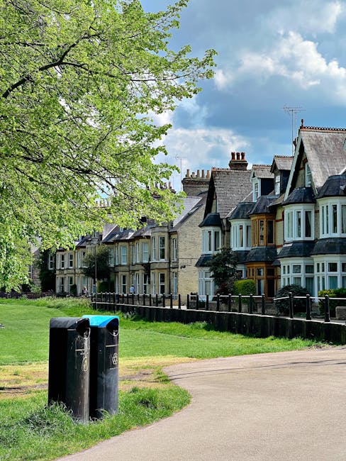 A residential street scene featuring a lush, green tree with fresh, light-green leaves on the left side of the image. In the foreground, there are two black and blue rubbish and recycling bins situated on a small patch of grass next to a paved footpath. Behind the bins, a row of traditional terraced houses with varying architectural styles is visible, each with multiple bay windows, some framed with white or wooden detailing, and pitched roofs with chimneys. The houses are constructed from light-colored brick and painted finishes, and are separated by a low black metal fence. The sky overhead is partly cloudy with patches of blue, creating a bright, natural lighting environment suitable for outdoor waste collection or private rubbish removal activities, with the scene reflecting typical urban residential areas where independent waste disposal methods might be employed by companies such as House Clearance Finchley.