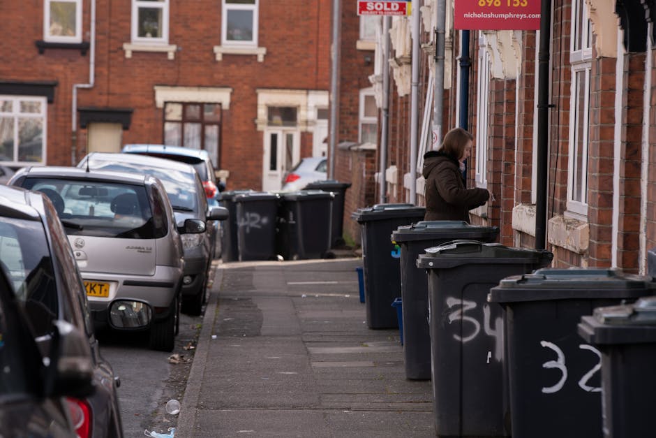 A woman standing on a narrow pavement next to a brick residential building, with her back slightly turned, near a row of large black wheelie bins used for rubbish disposal. The bins are lined up along the facade of the building, with some marked with white spray-painted numbers. In the background, there are several parked cars, including a grey hatchback closest to the woman, and additional residential buildings constructed with red brick and white window frames visible further along the street. The scene is illuminated by natural daylight, with a slight overcast sky, creating a neutral, professional atmosphere typical of urban rubbish collection areas. The presence of multiple bins and parked vehicles suggests this could be a designated collection or private waste handling zone often used in independent rubbish removal services, such as those offered by House Clearance Finchley, supporting the context of alternative waste management outside local authority collections.