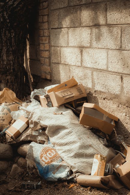 A pile of mixed waste and packaging materials, including cardboard boxes, paper bags, and plastic wrapping, is situated on the ground next to a rough stone and brick wall, with a tree trunk visible on the left side. The cardboard boxes vary in size, some partially crushed or torn, with visible printed markings and brown kraft finishes. A large, textured white sack or bag is amidst the collection, holding some of the waste. The scene appears to be an outdoor area, possibly an alleyway or back yard, where rubbish has been gathered for collection or disposal. The lighting suggests natural daylight, and the environment reflects a typical site for private waste clearance arrangements, such as those provided by companies like House Clearance Finchley, who specialise in rubbish removal services that support alternative waste handling outside of local authority collection. Overall, the scene underscores the practical setup for waste removal tasks involving loose and boxed refuse ready for collection or disposal.