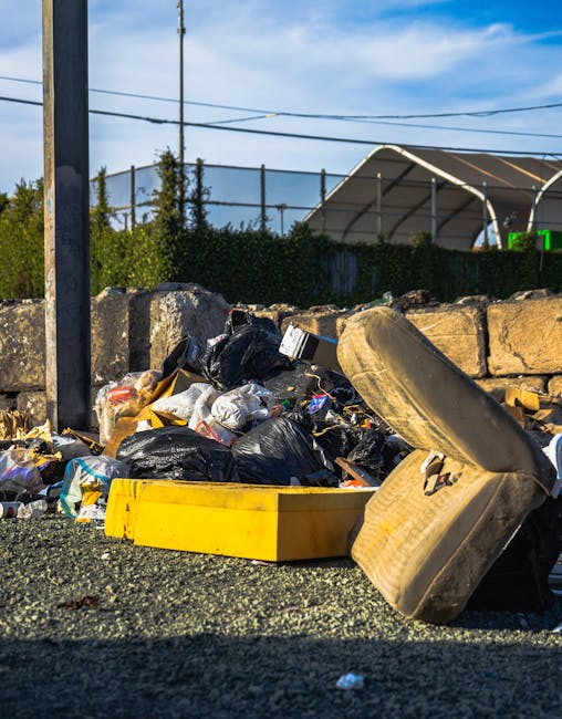 A pile of mixed waste and packaging materials, including cardboard boxes, paper bags, and plastic wrapping, is situated on the ground next to a rough stone and brick wall, with a tree trunk visible on the left side. The cardboard boxes vary in size, some partially crushed or torn, with visible printed markings and brown kraft finishes. A large, textured white sack or bag is amidst the collection, holding some of the waste. The scene appears to be an outdoor area, possibly an alleyway or back yard, where rubbish has been gathered for collection or disposal. The lighting suggests natural daylight, and the environment reflects a typical site for private waste clearance arrangements, such as those provided by companies like House Clearance Finchley, who specialise in rubbish removal services that support alternative waste handling outside of local authority collection. Overall, the scene underscores the practical setup for waste removal tasks involving loose and boxed refuse ready for collection or disposal.