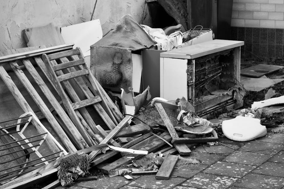 A pile of miscellaneous debris and discarded items situated on a paved outdoor area adjacent to a brick wall and a wooden fence. The collection includes a toppled wooden garden chair with slatted seats and backrest, showing weathered and worn textures, leaning against a larger piece of furniture or structure. There is a small, white plastic container or lid near the ground, alongside broken pieces of wood, possibly parts of furniture or shelving, and a section of old, damaged insulation or foam. An open-fronted appliance or cabinet, with visible shelving and some present internal components, sits partially obscured by the surrounding rubbish. Covering items are partly draped over the debris, suggesting temporary storage or a haphazard collection typical of on-site clearance. The scene displays typical waste materials often encountered during private rubbish removal or house clearance activities, emphasizing the type of clutter handled by services like House Clearance Finchley in the context of independent rubbish disposal. The natural lighting highlights the textures, contours, and variety of materials in the collection, illustrating a typical example of domestic or garden waste awaiting collection or disposal in an environmentally neutral setting.