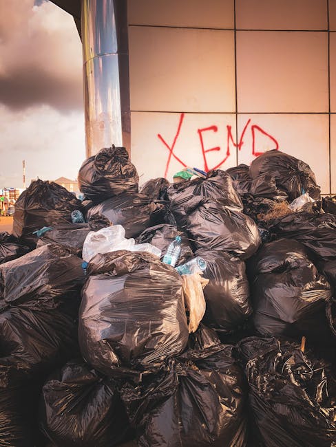 A large accumulation of black plastic rubbish bags stacked against a tiled wall, with some bags appearing to be overfilled and bulging. A few transparent bottles and plastic wrappers are visible protruding from the bags. The wall behind features a graffiti mark in red spray paint, spelling out 'XEND.' The scene is outdoors, with an overcast sky partially visible above, suggesting a city environment where private waste collection might be used due to limited space for waste disposal. The overall impression highlights the challenges faced in rubbish removal where narrow streets and restricted access complicate collection efforts, making the presence of bags and scattered waste a common sight in some urban alleyways or service areas. The image is relevant to the topic of independent rubbish clearance services, such as those offered by House Clearance Finchley, especially in situations requiring prompt removal of accumulated waste in tight or hard-to-reach areas.