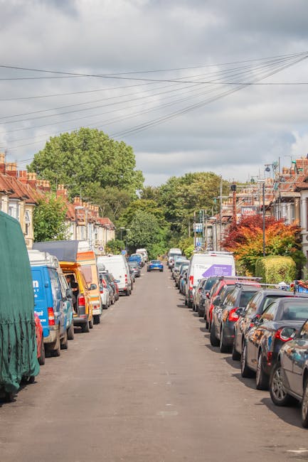 A residential street in Finchley with parked cars lining both sides of the narrow asphalt roadway, including a variety of sedans, hatchbacks, and a small van, some of which are covered with protective tarps. The driveway is bordered by rows of traditional brick terraced houses with tiled roofs, small front gardens, and some with visible chimneys. Pedestrian-facing street lamps and utility poles with overhead power lines run parallel to the pavement, while trees and greenery, including a prominent leafy tree on the left and a shrub with reddish foliage on the right, provide a natural backdrop. The sky overhead is partly cloudy with patches of sunlight filtering through, casting subtle shadows. This scene illustrates the challenges of access for private rubbish clearance services on narrow residential streets, where parking and street width may impact the logistics of waste removal, an area in which House Clearance Finchley offers alternative collections for rubbish management.