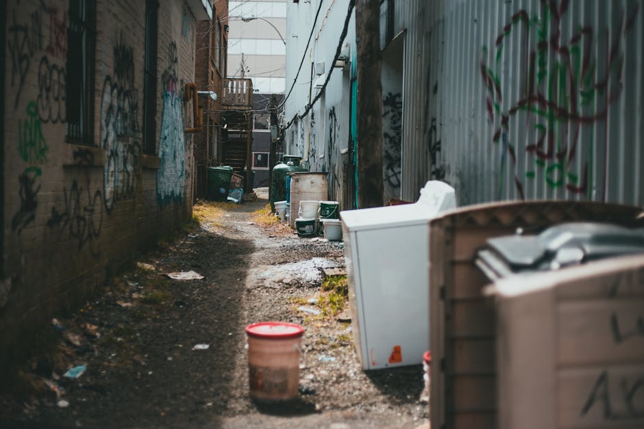 A large accumulation of black plastic rubbish bags stacked against a tiled wall, with some bags appearing to be overfilled and bulging. A few transparent bottles and plastic wrappers are visible protruding from the bags. The wall behind features a graffiti mark in red spray paint, spelling out 'XEND.' The scene is outdoors, with an overcast sky partially visible above, suggesting a city environment where private waste collection might be used due to limited space for waste disposal. The overall impression highlights the challenges faced in rubbish removal where narrow streets and restricted access complicate collection efforts, making the presence of bags and scattered waste a common sight in some urban alleyways or service areas. The image is relevant to the topic of independent rubbish clearance services, such as those offered by House Clearance Finchley, especially in situations requiring prompt removal of accumulated waste in tight or hard-to-reach areas.