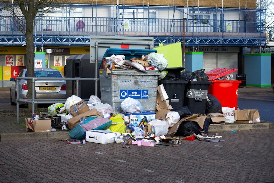 A public outdoor waste disposal area in a town center showing multiple overflowing rubbish bins and surrounding discarded waste materials. The scene includes a large gray bin labeled for mixed paper and card with open lid, revealing crumpled paper, cardboard boxes, and paper packaging sticking out. Next to it are black and black-and-gray bins overflowing with plastic bags filled with various rubbish, some spilling onto the paved ground. A red recycling bin is also visible, partially covered by black trash bags and loose debris. In front of the bins, various refuse including flattened cardboard boxes, plastic containers, paper packets, and assorted packaging are scattered across the asphalt surface. Behind the waste collection point, a building with a blue scaffolding structure and green panel accents is visible, with a storefront displaying signage for a fish bar and other small shops. The area appears to be in an urban setting, with a parked silver car and a low metal railing separating the waste site from the sidewalk, highlighting the importance of organized rubbish removal and proper waste disposal for maintaining cleanliness.