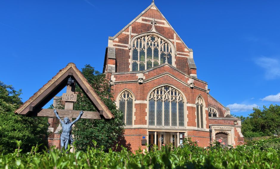 A large historic brick church with Gothic architectural features, including tall pointed arched windows with intricate tracery and stained glass, and a steeply pitched roof with a small spire at the top. The building's exterior showcases a combination of red and beige bricks, with decorative stone accents around the windows and roofline. In the foreground, there is a wooden crucifix shelter with a small triangular roof, featuring a detailed metal sculpture of Jesus Christ mounted on a cross, with the inscription 'INRI' above his head. The crucifix is positioned to the left side of the image, partially surrounded by lush green shrubbery and low plants that frame the scene at the bottom. The church is situated outdoors on a clear sunny day, with a bright blue sky and a few wispy clouds, and some trees are visible around the building, emphasizing its historic and churchyard setting. This scene is typical of a historic place of worship with a nearby graveyard or garden area, often associated with independent or private waste handling services like those offered by House Clearance Finchley.
