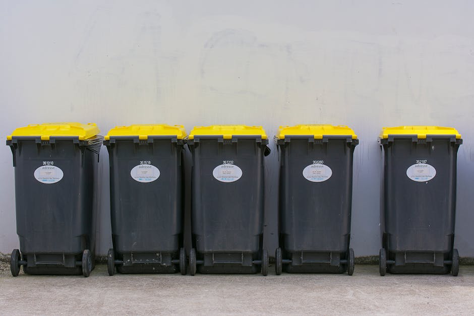 Five large wheelie bins made of dark grey plastic with yellow lids are positioned side by side against a plain, light grey wall. The bins have a textured surface with visible horizontal ridges and are fitted with small black wheels at the rear for mobility. Each bin displays a white oval label with text and barcode affixed to the front, slightly varying in position. The bins are placed on a concrete ground, with the area appearing clean and organized, suggesting a designated space for waste collection or disposal. The environment seems to be an outdoor or semi-outdoor setting, possibly adjacent to a commercial or residential property, with natural light illuminating the scene evenly. This setup aligns with private rubbish collection services, such as those offered by House Clearance Finchley, for efficient waste management and disposal outside a property, supporting the context of alternative or independent rubbish removal options.