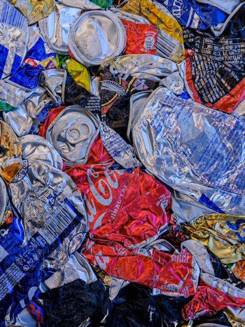 A large outdoor collection of waste and rubbish is piled up on a paved urban street pavement in front of a commercial building. The accumulation includes various types of waste, such as cardboard boxes, plastic bags, paper debris, and other miscellaneous refuse. In the foreground, numerous flattened and crumpled cardboard boxes, some with printed logos or labels, are scattered across the ground and partially spilling onto the pavement. Several black and colored plastic garbage bags are stacked next to a large, gray recycling bin marked for mixed paper and cardboard, with its lid open and waste overflowing. Adjacent to the recycling bin, there are larger black and red waste containers, some with black garbage bags protruding. Behind the waste, a blue roller shutter door is visible, indicating a commercial space, with signage and protective fencing on the upper part of the building. To the left, a silver car is parked behind a metal railing, and a tree trunk extends upward out of the frame. The environment suggests a typical urban scene awaiting rubbish collection, and the scene supports the context of private waste handling or alternative rubbish removal services like those provided by House Clearance Finchley.